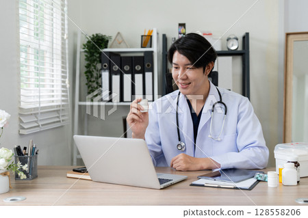 Young Asian Male Doctor Conducting Online Consultation in Modern Office with Laptop and Medical Equipment Young Asian Male Doctor Conducting Online Consultation in Modern Office with Laptop and Medical Equipment 128558206