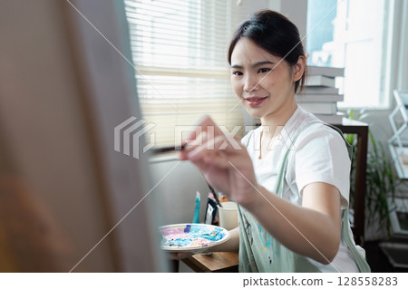 Young Woman Artist Painting on Canvas in Bright Studio with Natural Light Young Woman Artist Painting on Canvas in Bright Studio with Natural Light 128558283