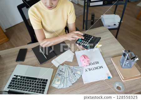 Young Woman Calculating Monthly Expenses at Home Office Desk with Calculator, Cash, and Financial Documents Young Woman Calculating Monthly Expenses at Home Office Desk with Calculator, Cash, and Financial Documents 128558325