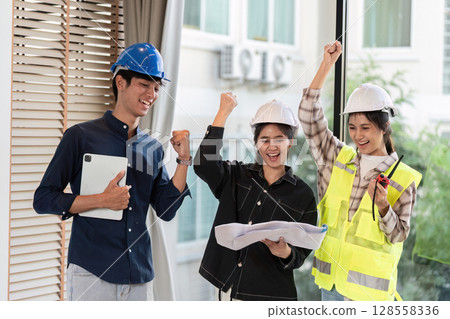 Group of Happy Engineers Celebrating Success at Construction Site with Blueprints and Safety Gear 128558336