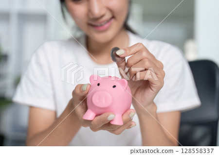 Woman Saving Money by Putting Coin into Pink Piggy Bank, Financial Planning and Savings Concept 128558337