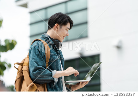 Young Man Using Laptop Outdoors with Modern Building in Background, Wearing Denim Jacket and Backpack Young Man Using Laptop Outdoors with Modern Building in Background, Wearing Denim Jacket and Backpack 128558357