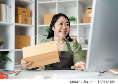 Smiling Woman Holding Package While Working on Computer in Home Office with Shelves in Background 128558362