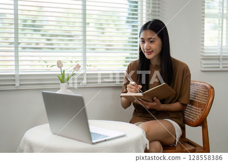 Young Woman Working from Home with Laptop and Notebook in Bright Modern Living Room 128558386