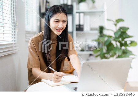 Young Woman Using Laptop and Taking Notes in Modern Home Office with Green Plants and Natural Light Young Woman Using Laptop and Taking Notes in Modern Home Office with Green Plants and Natural Light 128558440