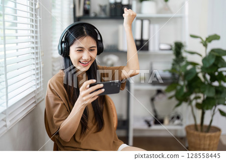Young Woman Enjoying Mobile Gaming at Home with Headphones, Celebrating Victory in a Modern Living Room 128558445