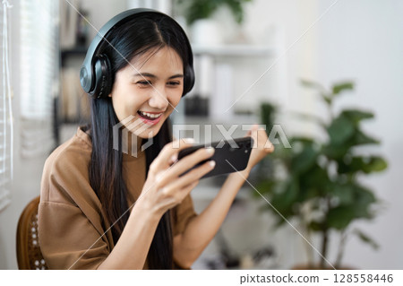Young Woman Enjoying Mobile Gaming with Headphones in Modern Home Environment 128558446
