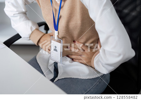 Person Experiencing Stomach Pain at Work, Holding Stomach in Office Chair, Wearing ID Badge Person Experiencing Stomach Pain at Work, Holding Stomach in Office Chair, Wearing ID Badge 128558482