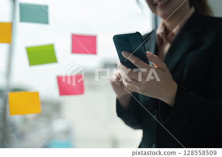 Professional Businesswoman Using Smartphone in Modern Office with Colorful Sticky Notes on Glass Wall Professional Businesswoman Using Smartphone in Modern Office with Colorful Sticky Notes on Glass Wall 128558509