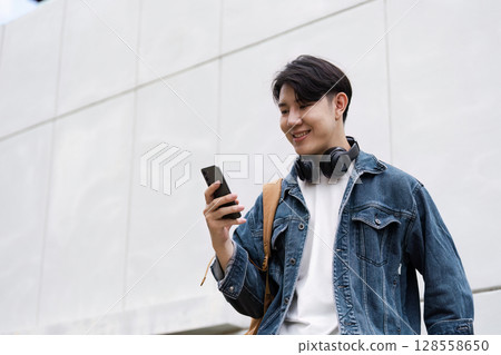 Young Student Smiling While Using Smartphone Outdoors, Wearing Headphones and Backpack, Casual Denim Jacket 128558650
