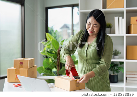 Small Business Owner Packing Products for Shipping in Home Office with Green Plants and Shelves in Background 128558889