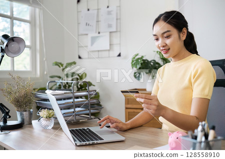 Young Woman Using Credit Card for Online Banking on Laptop at Home Office Desk with Modern Decor Young Woman Using Credit Card for Online Banking on Laptop at Home Office Desk with Modern Decor 128558910