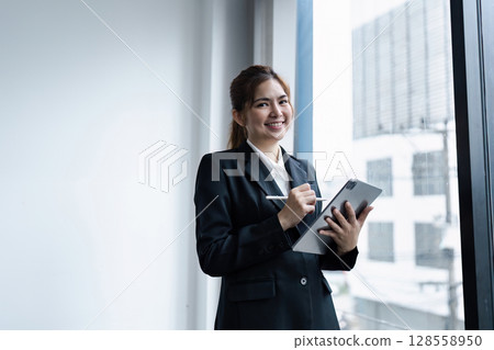 Confident Businesswoman in Modern Office Holding Tablet and Smiling by Window 128558950