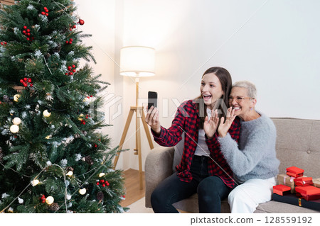 Grandmother and Granddaughter Video Calling Family on Christmas Morning by the Decorated Tree Grandmother and Granddaughter Video Calling Family on Christmas Morning by the Decorated Tree 128559192