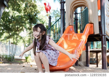 Happy Young Girl Sliding Down Playground Slide on a Sunny Day in the Park Happy Young Girl Sliding Down Playground Slide on a Sunny Day in the Park 128559193
