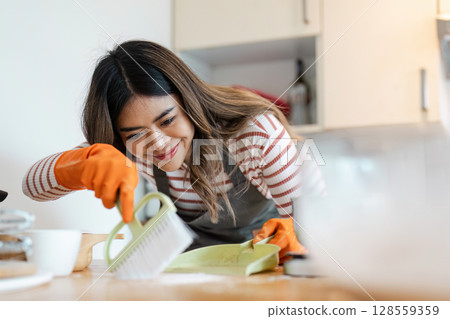 Woman Cleaning Kitchen Counter with Brush and Dustpan, Wearing Orange Gloves, Smiling and Focused on Household Chores 128559359