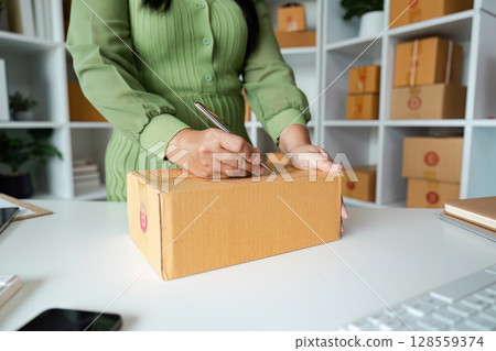 Small Business Owner Preparing Package for Shipment in Modern Office Space with Shelves of Boxes Small Business Owner Preparing Package for Shipment in Modern Office Space with Shelves of Boxes 128559374