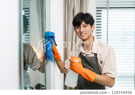 Young Man Cleaning Window with Spray Bottle and Cloth, Wearing Apron and Gloves, Smiling at Camera, Modern Home Interior 128559456