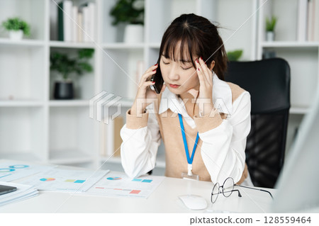 Stressed Businesswoman on Phone Call in Modern Office with Documents and Charts on Desk 128559464