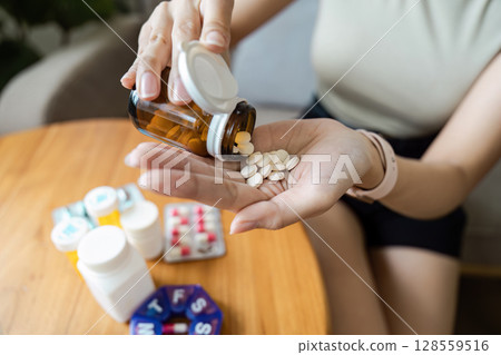 Close-up of a Person Pouring Pills from a Bottle into Their Hand with Various Medications on a Wooden Table 128559516