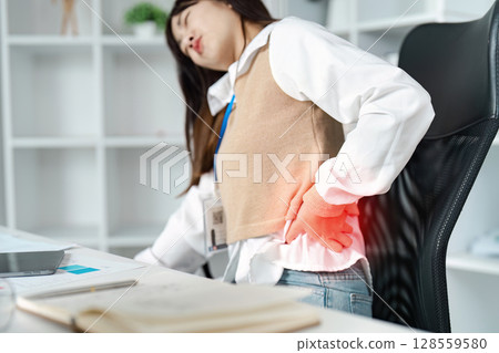 Young Woman Experiencing Severe Back Pain While Working at Desk in Modern Office Setting 128559580