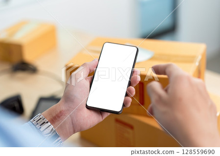 E-commerce management. A man pointing at smartphone screen while managing packages. E-commerce management. A man pointing at smartphone screen while managing packages. 128559690
