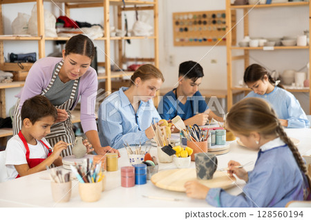 Children paint ceramic work-pieces at table with teacher in art studio 128560194