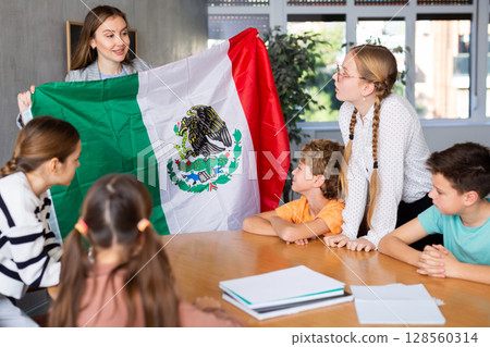 Smiling young female teacher conducting lesson for preteens, showing state flag of Mexico in audience of middle school 128560314
