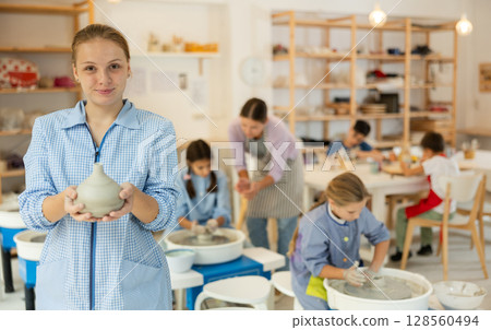 Girl holds a clay product against the background of students and a teacher 128560494