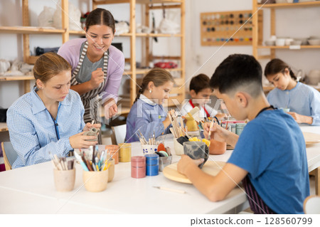 Children paint ceramic work-pieces at table with teacher in art studio 128560799