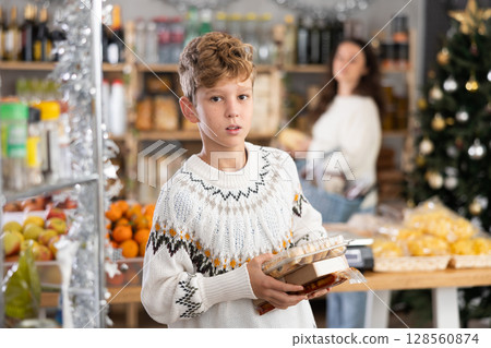 Teen boy selects sweets near window case in store, hold some package, New Year Eve 128560874