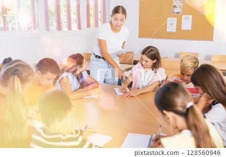 Female teacher giving lesson for preteen schoolchildren sitting around common desk 128560946