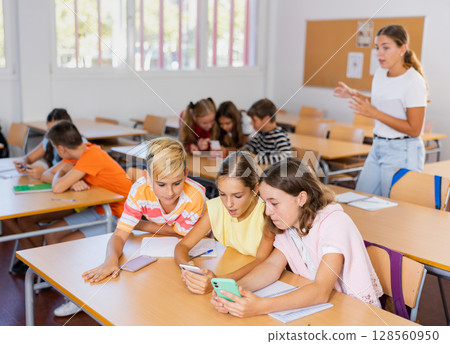 Modern curious preteen schoolchildren using smartphones at lesson Modern curious preteen schoolchildren using smartphones at lesson 128560950