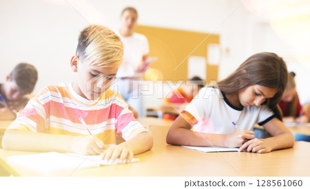 Diligent tweens listening to lecture of teacher and writing in notebooks 128561060