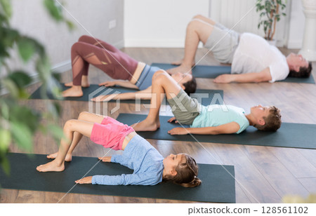 Teenage girl and boy with mom and dad do exercises lying on their backs in modern yoga studio Teenage girl and boy with mom and dad do exercises lying on their backs in modern yoga studio 128561102
