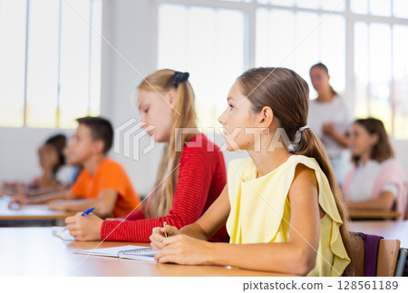 Interested preteen girl studying in classroom with classmates Interested preteen girl studying in classroom with classmates 128561189