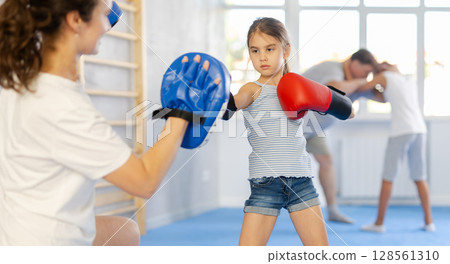 Tween girl in boxing gloves practicing punches on focus mitts in hands of mother 128561310