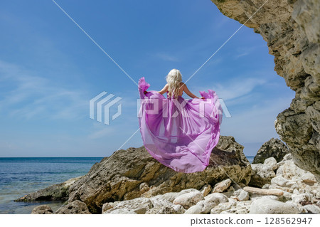 Dress ocean woman: Blonde poses in purple dress, on rocks by sea, sunny day for photoshoot. 128562947