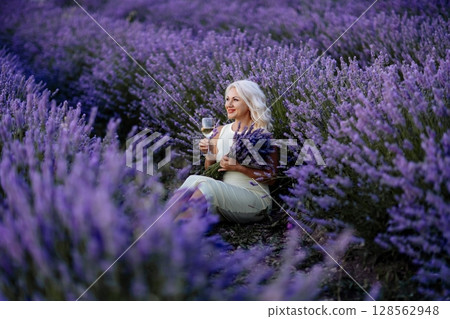 Blonde lavender field holds a glass of white wine in her hands. Happy woman in white dress enjoys lavender field picnic holding a large bouquet of lavender in her hands . Illustrating woman's picnic 128562948