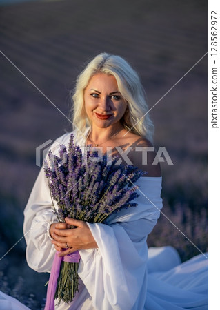 Blonde woman poses in lavender field at sunset. Happy woman in white dress holds lavender bouquet. Aromatherapy concept, lavender oil, photo session in lavender 128562972