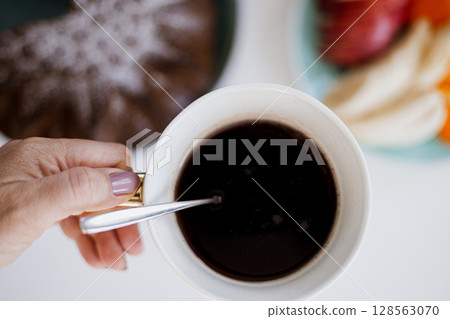 Close-up of a hand holding a coffee cup with cake and fruit on a bright breakfast table Close-up of a hand holding a coffee cup with cake and fruit on a bright breakfast table 128563070