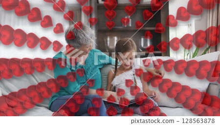 Image of hearts over happy caucasian grandmother and granddaughter reading book on sofa 128563878