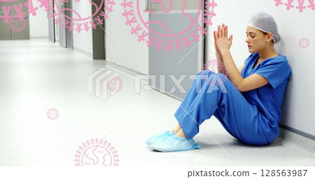 Image of pink globe and people logos, over distressed female surgeon in hospital corridor Image of pink globe and people logos, over distressed female surgeon in hospital corridor 128563987