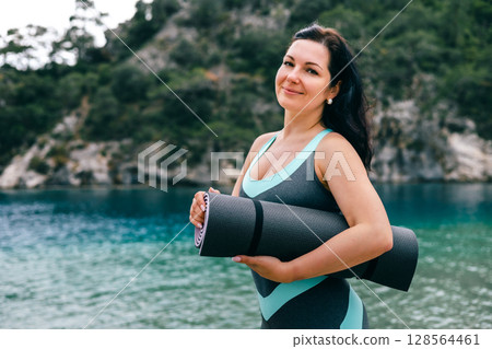 Smiling woman holding yoga mat by turquoise sea, ready for fitness session in nature 128564461