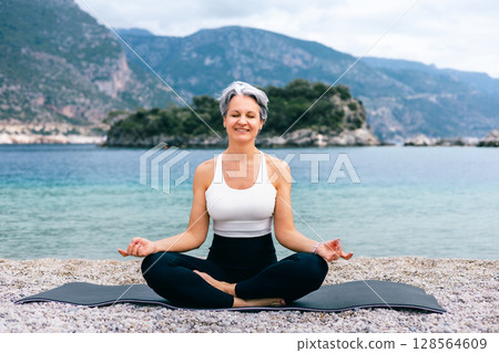 Senior woman meditating on beach with mountains in background, enjoying peaceful nature 128564609