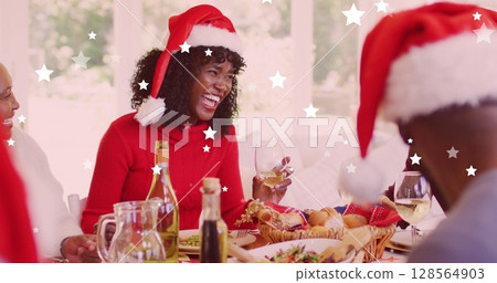 Image of stars falling over happy african american family wearing santa hats and having dinner 128564903
