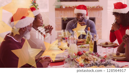 Image of stars falling over happy african american family wearing santa hats and praying 128564917