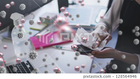Image of covid 19 cells floating over woman wiping smartphone in office 128564946
