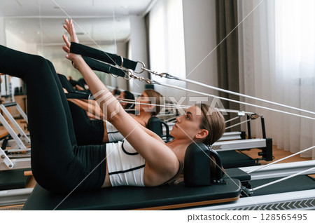 Side view. Stretching the legs. Women in fitness center are doing exercises Side view. Stretching the legs. Women in fitness center are doing exercises 128565495