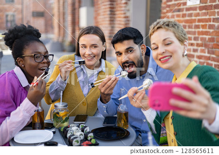 Friends enjoy sushi together, taking a selfie outdoors. They are smiling and having fun. 128565611